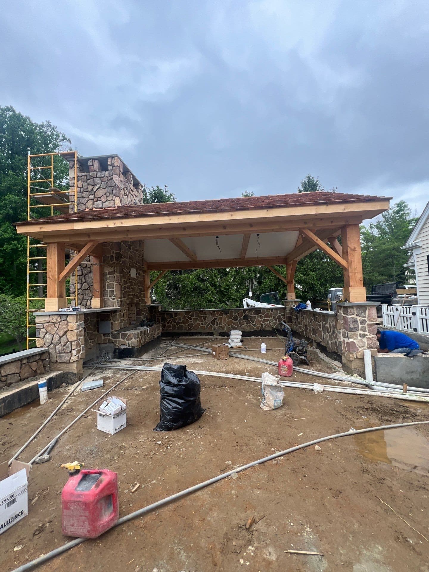 Cedar timber-frame pavilion under construction with a cedar-shake roof and a fieldstone fireplace and chimney.