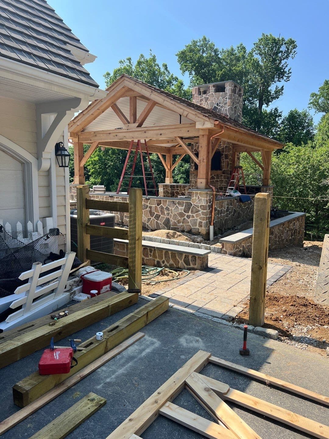Cedar timber-frame pavilion with a gable roof, king-post truss, and a fieldstone fireplace and chimney beside a home, on a paver patio.