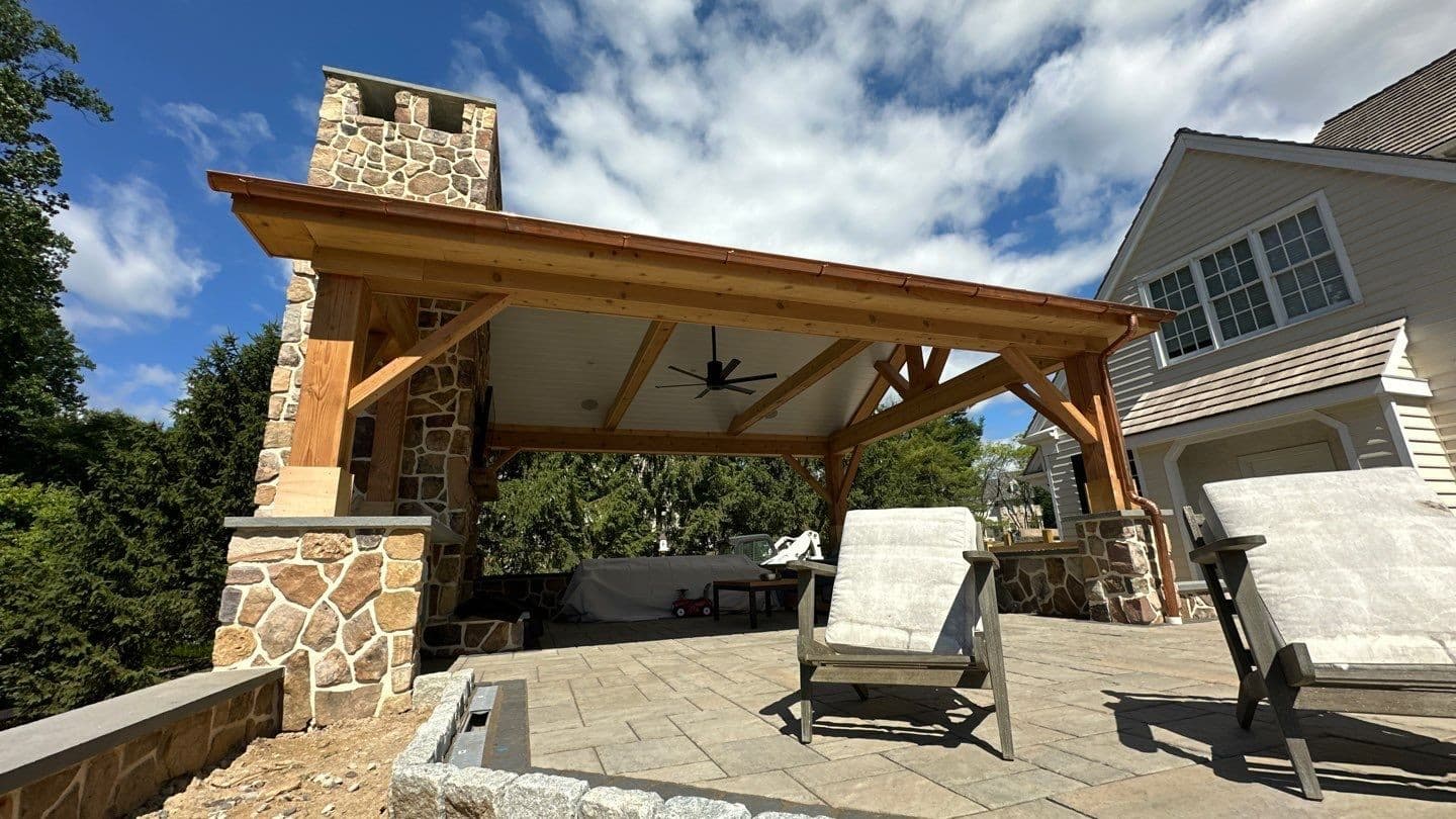 Cedar pavilion with a single-slope roof and a tall stone fireplace and chimney over a paver patio with Adirondack chairs beside a home.
