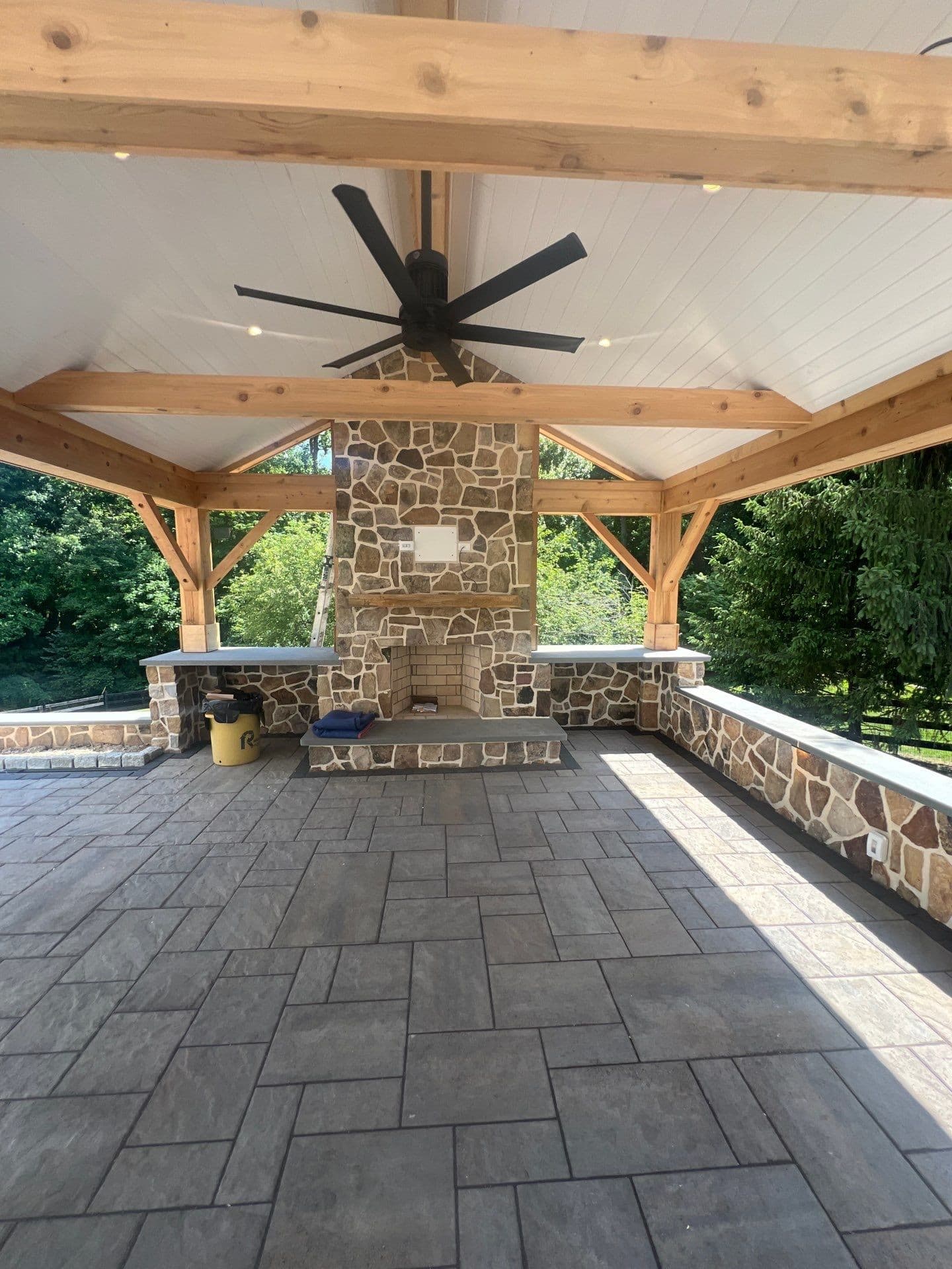 Interior of a cedar timber-frame pavilion with a vaulted tongue-and-groove ceiling, oversized black ceiling fan, and a floor-to-ceiling stone fireplace over a stamped-concrete patio.