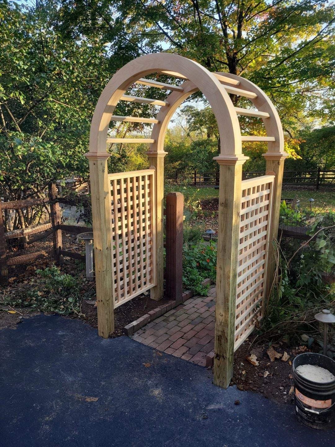 Cedar garden arbor with an arched top and lattice side panels set on a paver landing in a garden.