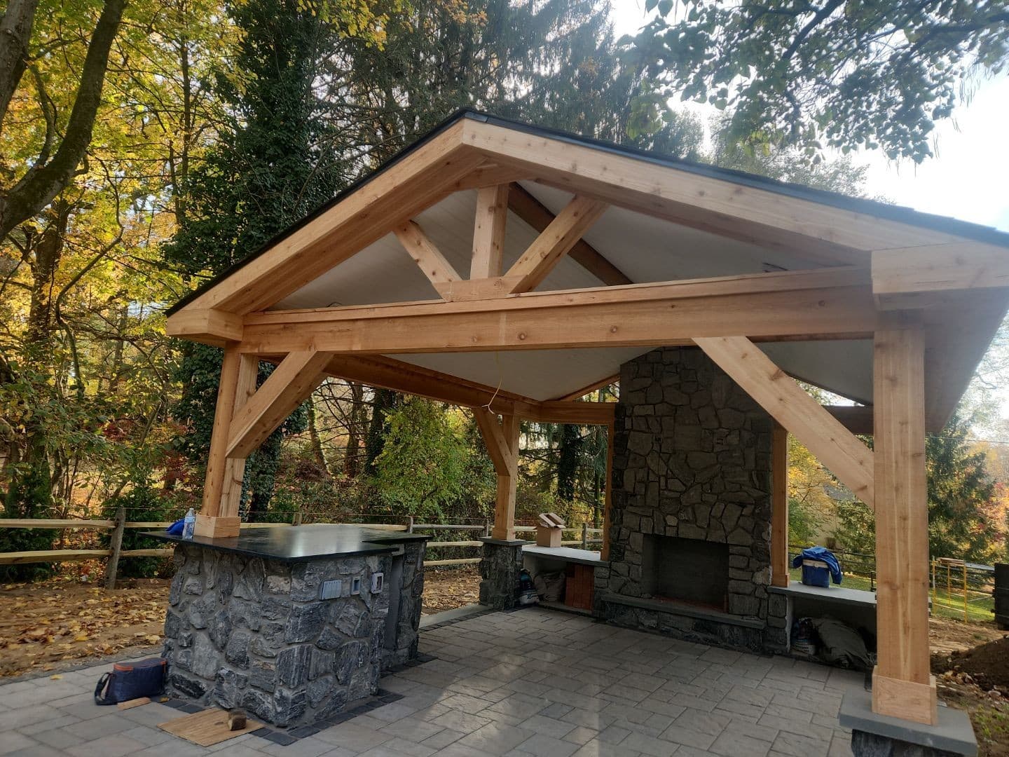 Cedar timber-frame pavilion with king-post trusses, a stone fireplace, and a stone outdoor-kitchen island with bluestone counters on a paver patio in an autumn setting.