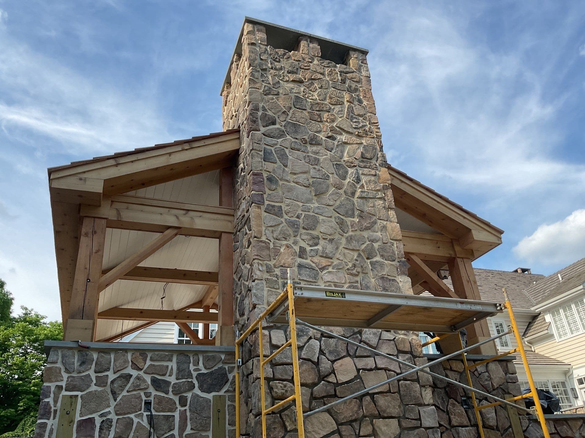 Tall fieldstone chimney of a cedar pavilion against a blue sky during construction.