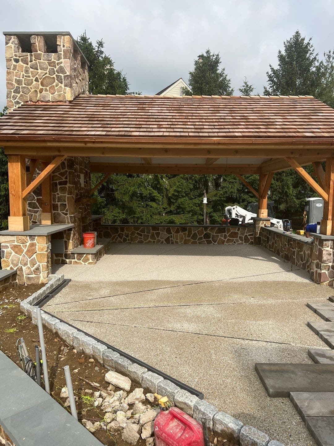 Cedar timber-frame pavilion with a cedar-shake roof, fieldstone fireplace and chimney, and stone half-walls over a concrete patio.