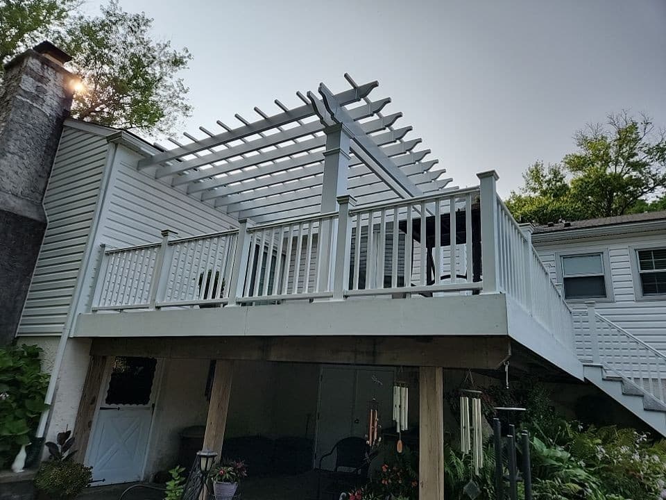 White vinyl pergola with an open rafter roof mounted on a raised deck with matching white railings, attached to a home.